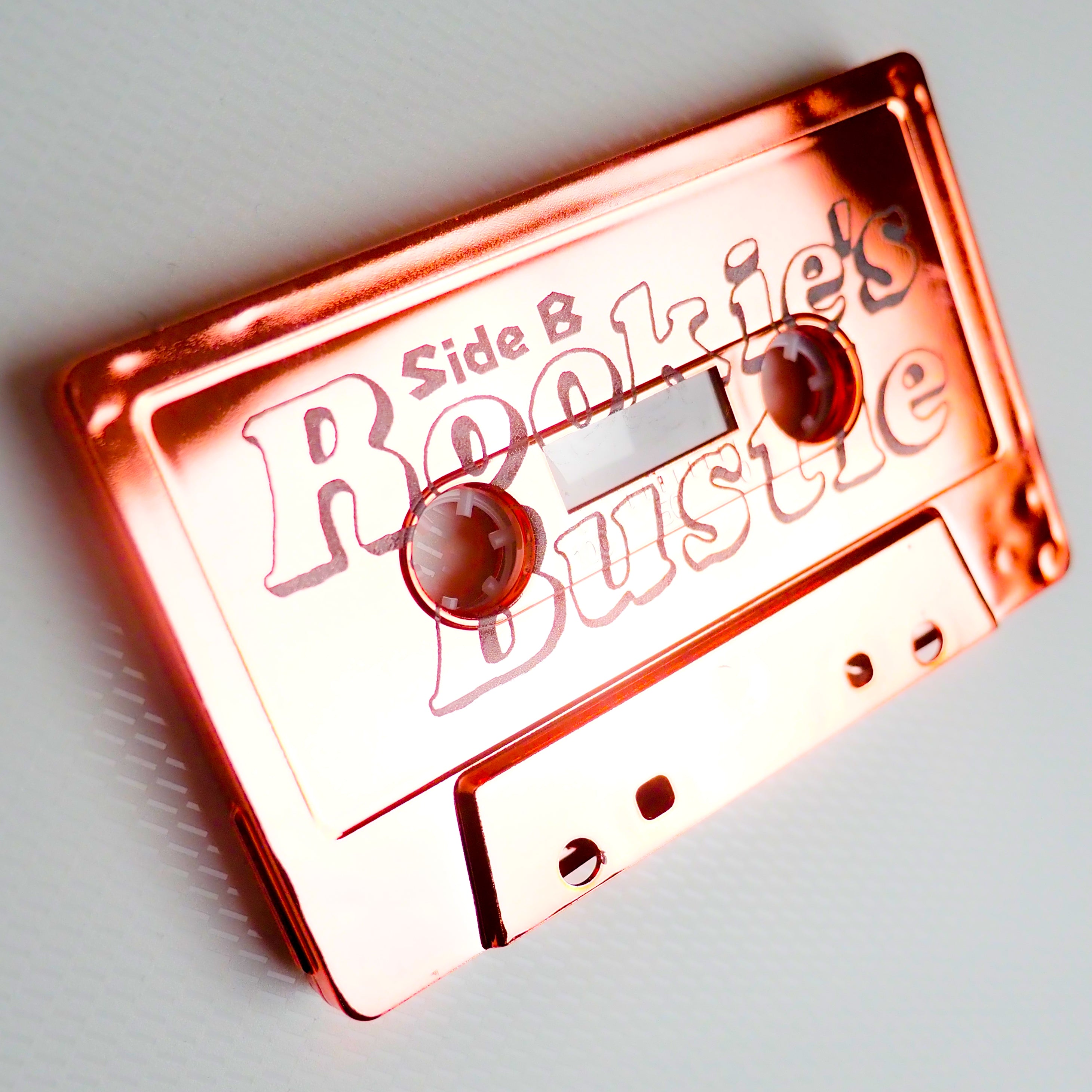 Rose gold cassette tape alone with 'Side B Rookie's Bustle' text on a white background