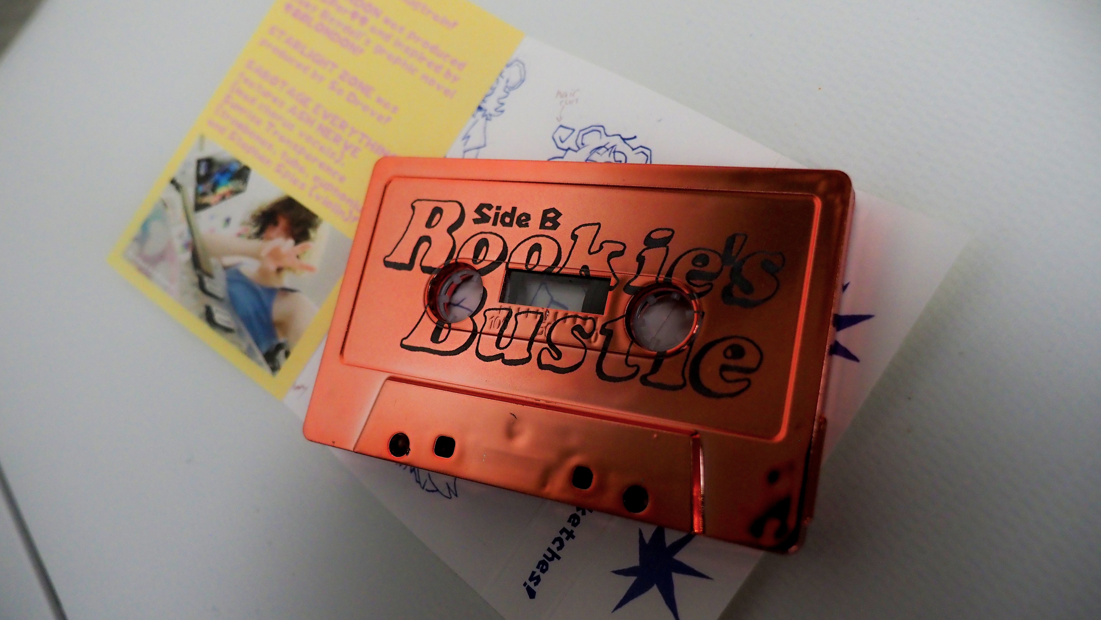 Orange cassette tape labeled 'Side B Rookie's Bustle' on a white surface with a yellow inset card.